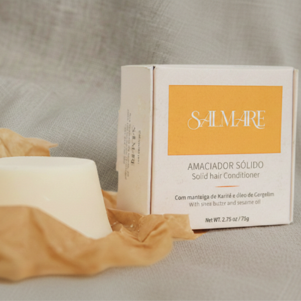 Amaciador Sólido Salmare redondo fora da embalagem branca e amarela, pousado sobre tecido de linho bege natural. Produto artesanal com Manteiga de Karité e Óleo de Gergelim visível na caixa.
