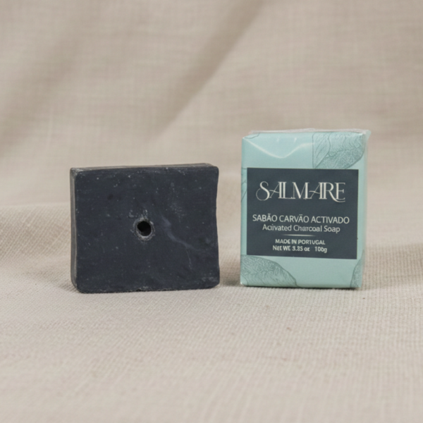 Sabão de Carvão Salmare barra redonda preta fora da embalagem branca com rótulo cinzento escuro, pousado sobre tecido de linho bege natural. Contém Carvão Ativado e Tea Tree.
