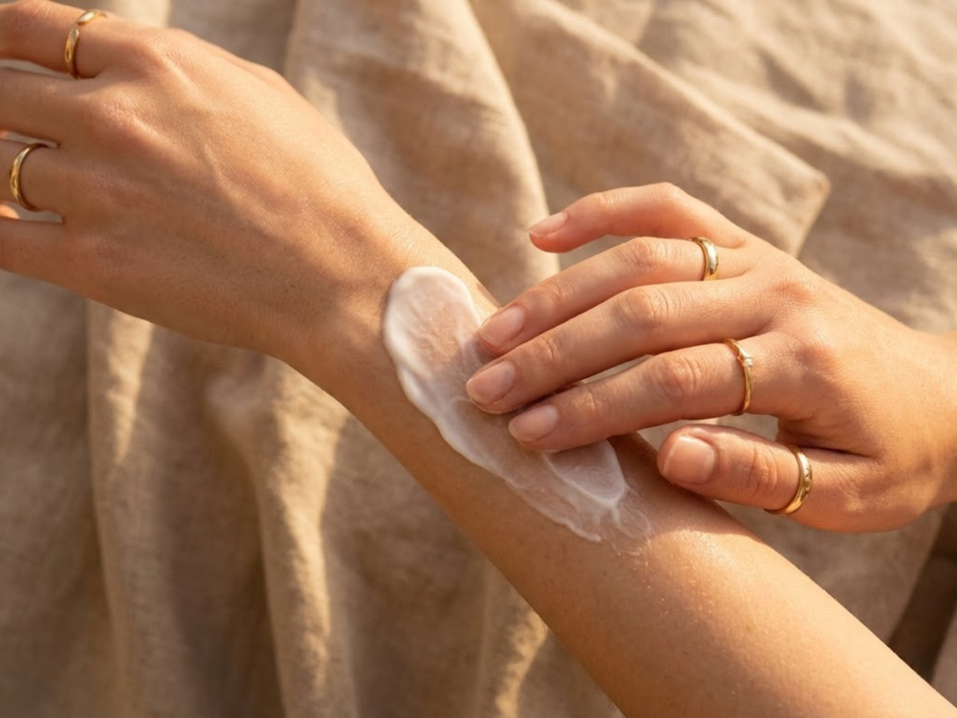 Close-up de mãos delicadas a massajar um creme hidratante rico na pele do braço, mostrando a textura suave e a absorção do produto. Iluminação solar dourada e fundo de linho bege desfocado.